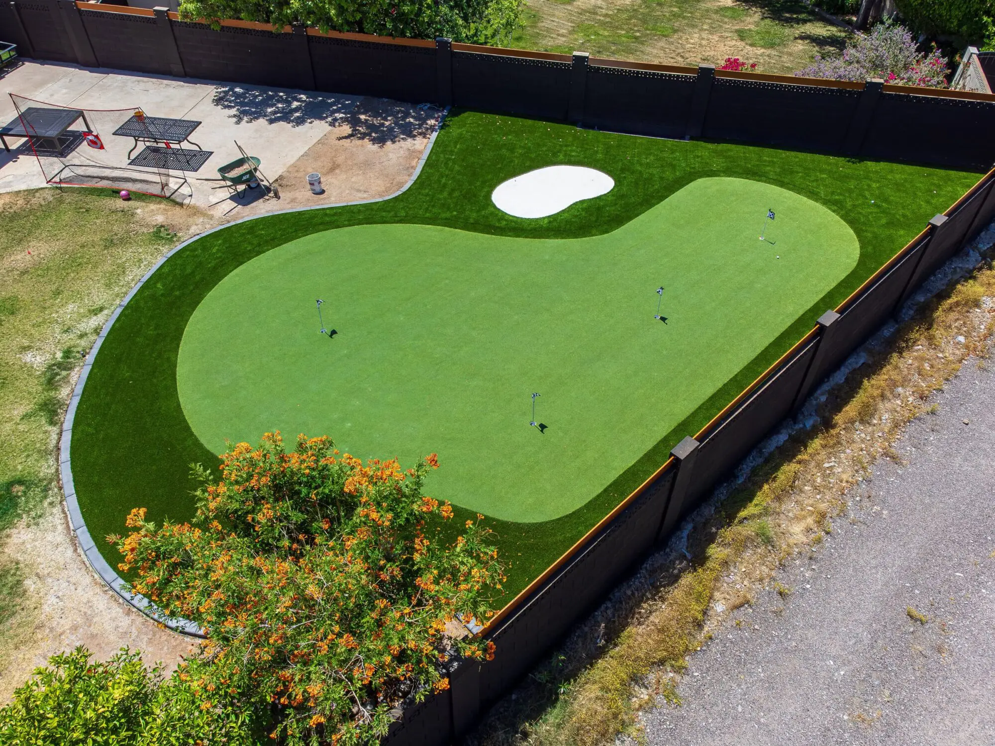 Aerial view of a backyard featuring a putting green nestled in artificial grass, complete with a small sand trap. Nearby, patio tables and chairs rest on a concrete area, all enclosed by a wooden fence.