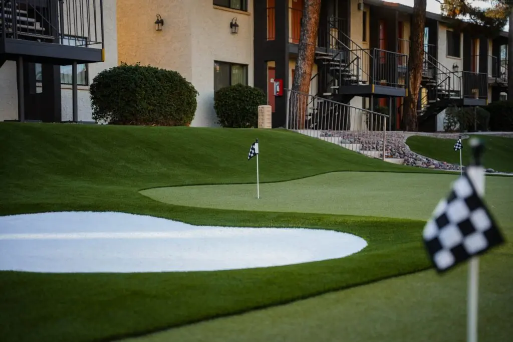 The well-manicured putting green features checkered flags and is encircled by neatly trimmed bushes. White sand traps accentuate the scene. In the background, a row of beige buildings with windows and exterior staircases completes this tranquil golf course setting.