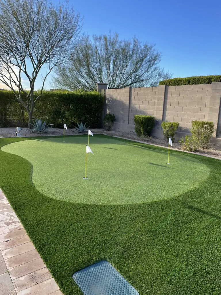 A backyard scene showcases a small putting green with three flagged holes on artificial turf, bordered by desert plants and a brick wall, all under a clear blue sky.