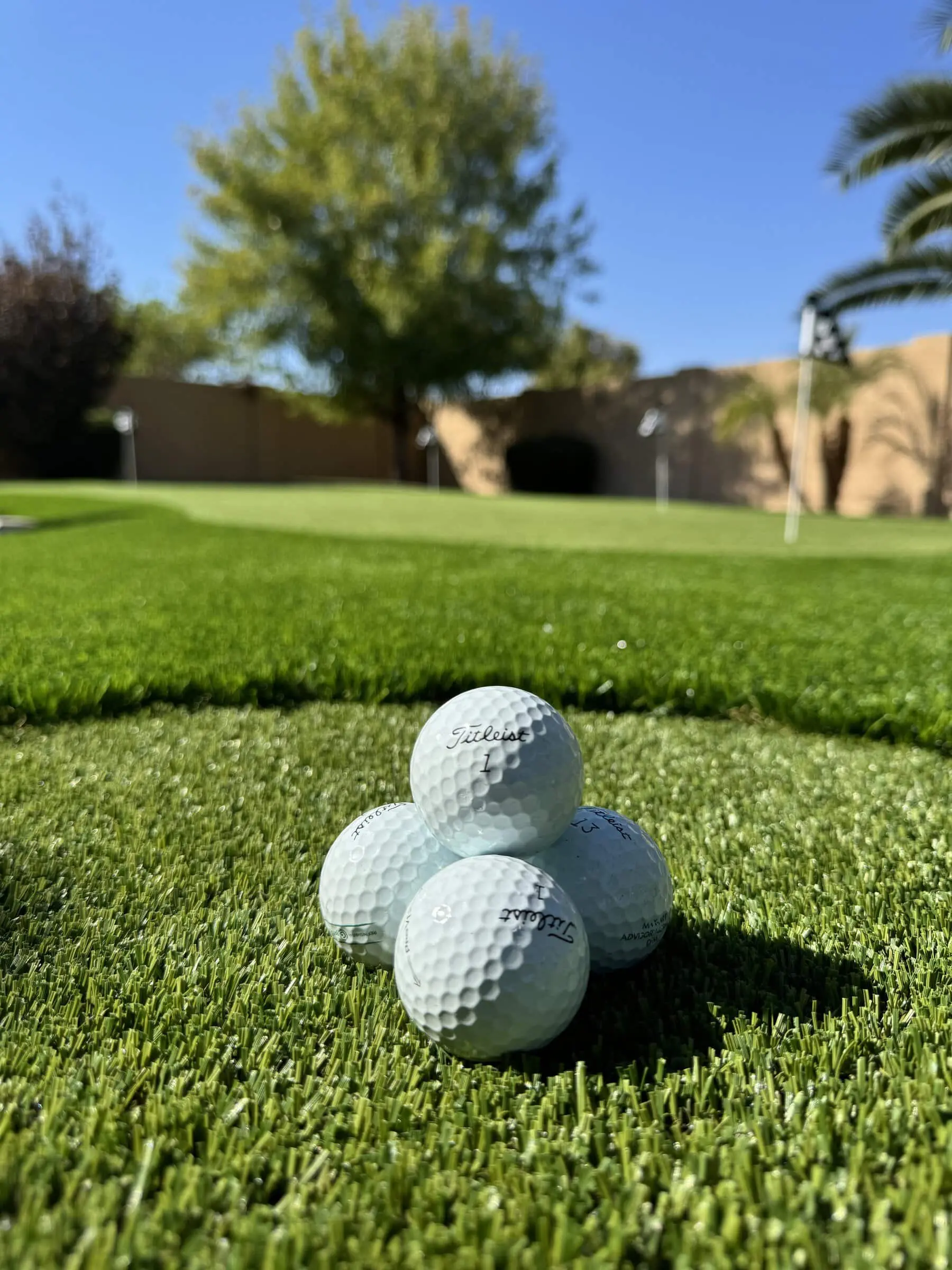 Four golf balls are stacked in a pyramid on lush green grass, perfectly poised near the putting green. In the background, trees stretch under a vibrant blue sky, capturing the essence of a sunny day on the golf course.