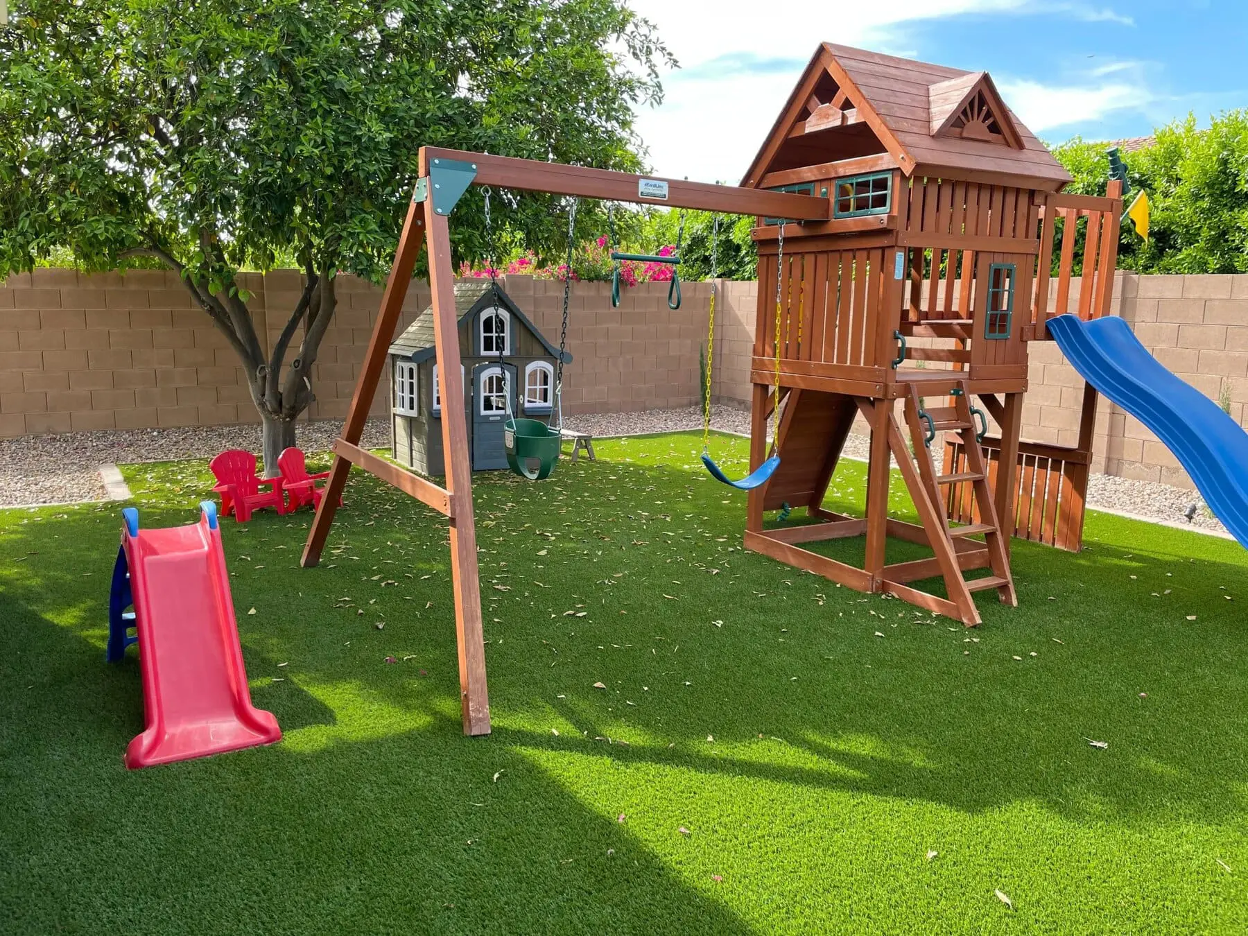 A backyard playground on Apex Turf features a wooden playset with a slide, swings, and climbing area. A small plastic slide and toy house are nearby, with trees and a brick wall in the background.