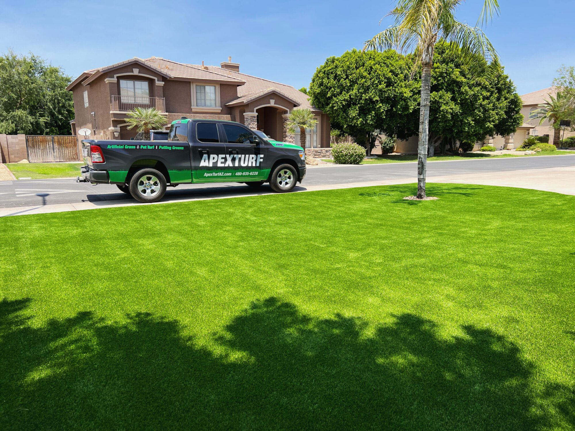A vibrant green artificial grass lawn is in the foreground with a pickup truck parked on the street. The truck, showcasing "APEX TURF" branding, hints at their expertise in artificial grass for Arizona. Suburban houses, trees, and a clear blue sky complete this idyllic scene.