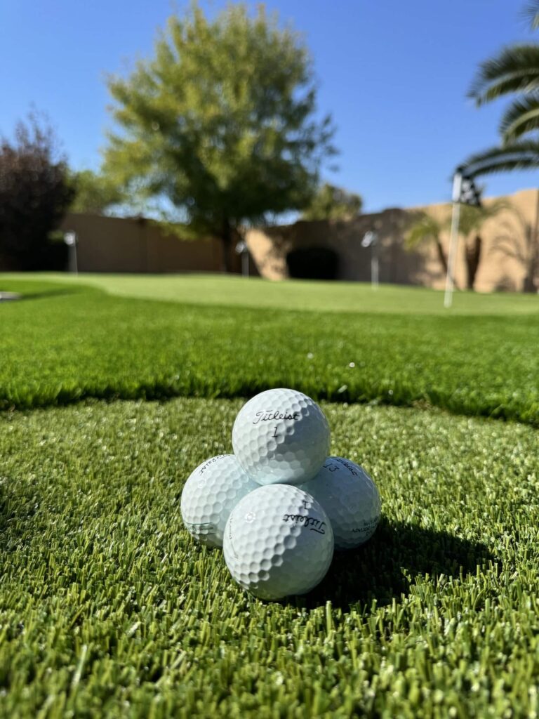 Four golf balls are stacked in a pyramid on lush green grass, perfectly poised near the putting green. In the background, trees stretch under a vibrant blue sky, capturing the essence of a sunny day on the golf course.