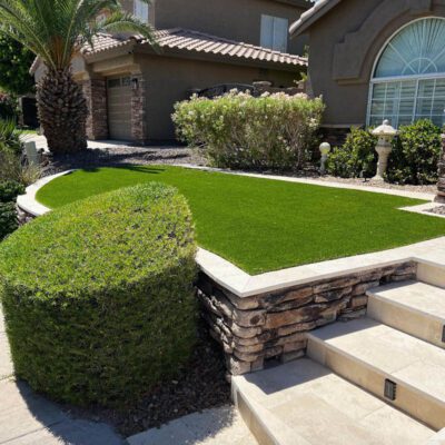 A well-maintained garden in Chandler features a rectangular patch of artificial grass by Apex Turf, elevated by stone walls. Surrounding shrubs and palm trees complement the tan, stone-accented house with arched windows. Stairs lead up to the entrance.