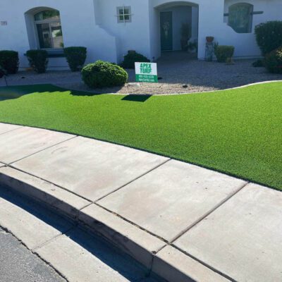 A suburban house with a well-manicured artificial grass lawn from Apex Turf, bordered by a concrete sidewalk. Shrubs decorate the landscape, and a sign reads 