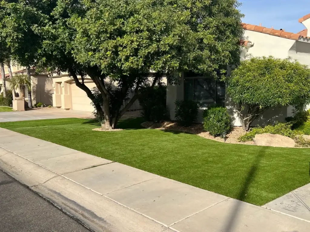 A suburban house with a well-maintained lawn and a large tree in the front yard. The house has a light exterior, a driveway, and shrubs near the windows. The pavement runs alongside the street.