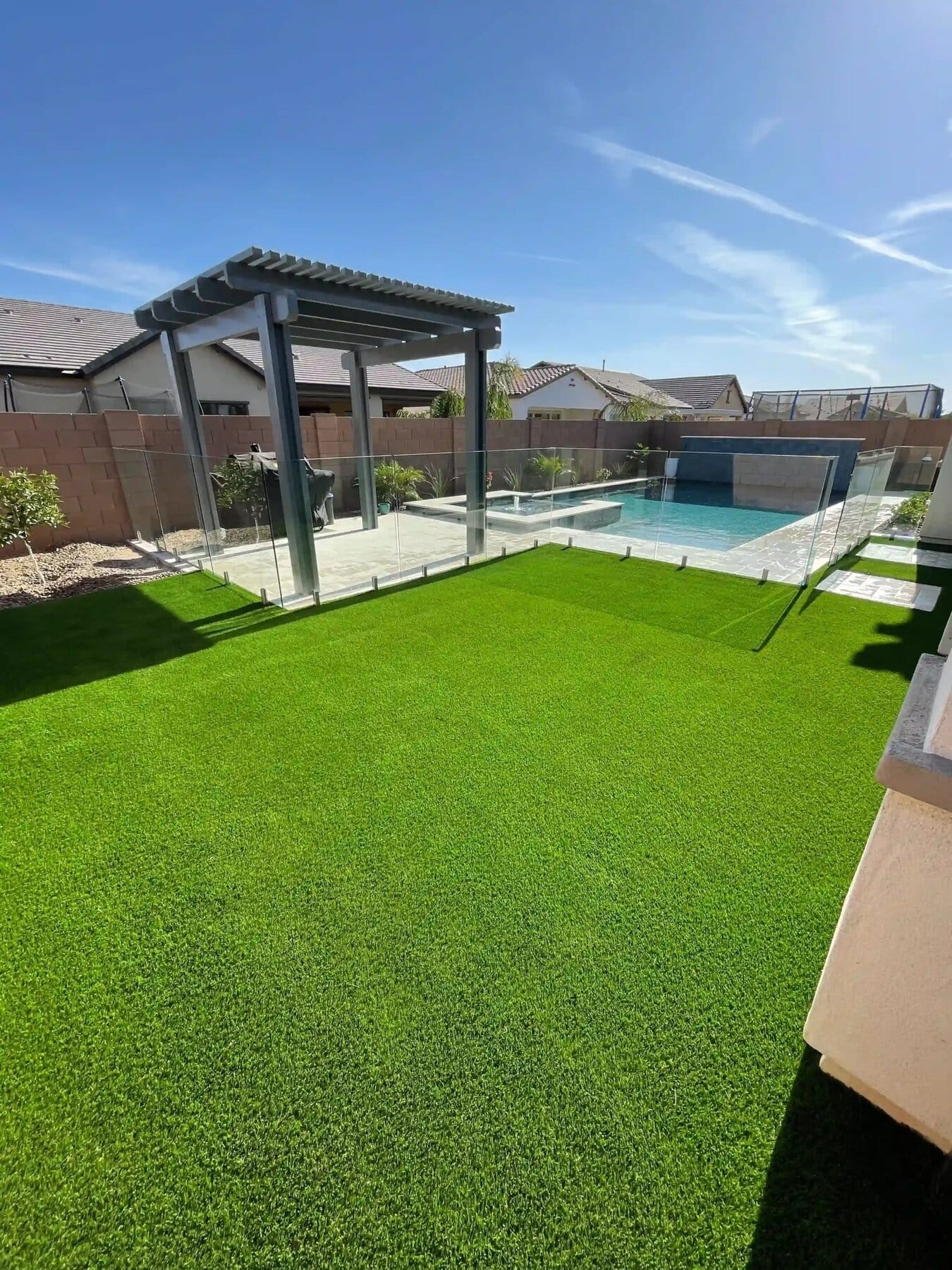 Manicured backyard featuring lush green Apex Turf, a modern pergola shading the dining area, and a glass-fenced swimming pool. Clear skies set the scene, with neighboring houses in view.