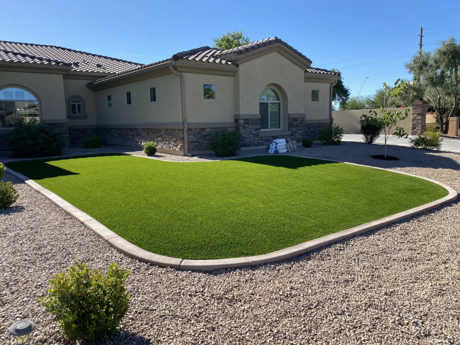 A modern house with a light beige exterior and arched windows sits behind a neatly landscaped yard. The yard boasts Apex Turf's bright green artificial grass bordered by a paver edge, surrounded by gravel and small bushes under a clear blue sky.