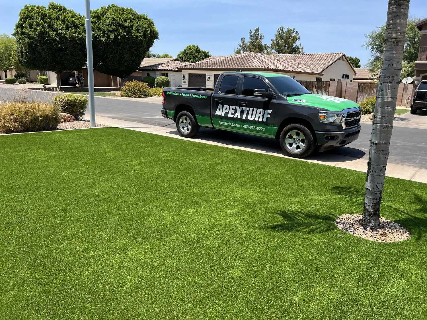A lush, green artificial grass lawn in a residential area. A green truck parked by the curb displays the word "Apex Turf." Trees, bushes, and houses surround the area under a clear sky.