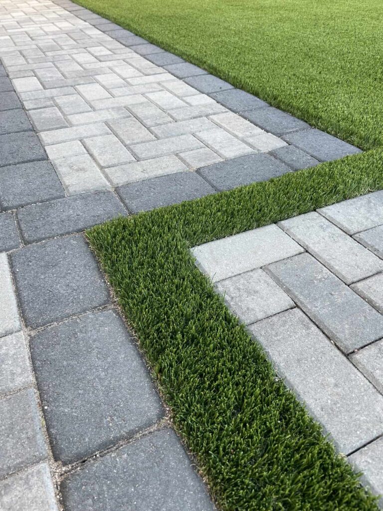 Grey concrete pavers laid in a herringbone pattern, with a dark grey paver border. Pavers installed next to artificial grass lawn