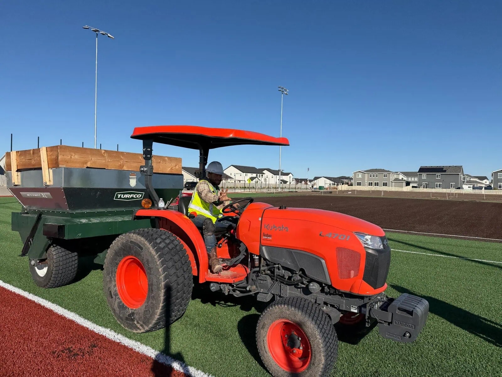 Image is of an Apex Turf staff member driving a tractor with the infill trailer, getting ready to distribute the rubber sports infill on the field