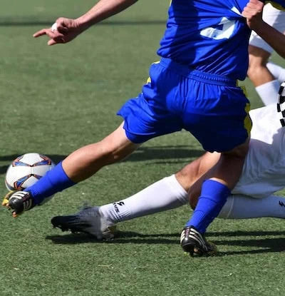Two soccer players with shin guards going after a soccer ball in a game. Photo taken from the waist down